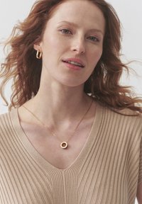 Pendientes de aro de oro y un collar con colgante de oro en una mujer con cabello rojo ondulado, que lleva un suéter de canalé en color beige y con cuello en V.