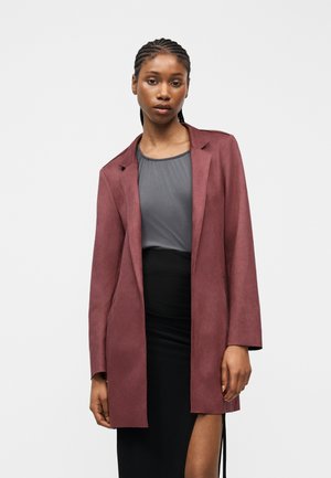 Femeie cu păr împletit, purtând un sacou burgund peste un top gri și o fustă neagră cu o tăietură pe coapsă, stând în fața unui fundal alb simplu.