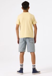 T-shirt jaune à manches courtes, shorts en denim bleu clair, chaussettes noires et blanches à motifs, et baskets montantes bleues.