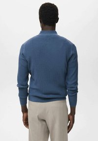 Ribana pulover v srednji modri barvi z ovratnikom, prilegajočimi se manšetami in pasom. Nosen preko bež hlač, kar poudarja sproščen dizajn.