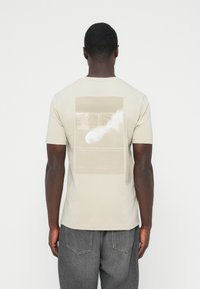 Béžna bombažna majica s kratkimi rokavi, na zadnji strani z otipljivim grafičnim potiskom v nevtralnih odtenkih. Gladek material, sproščen kroj.