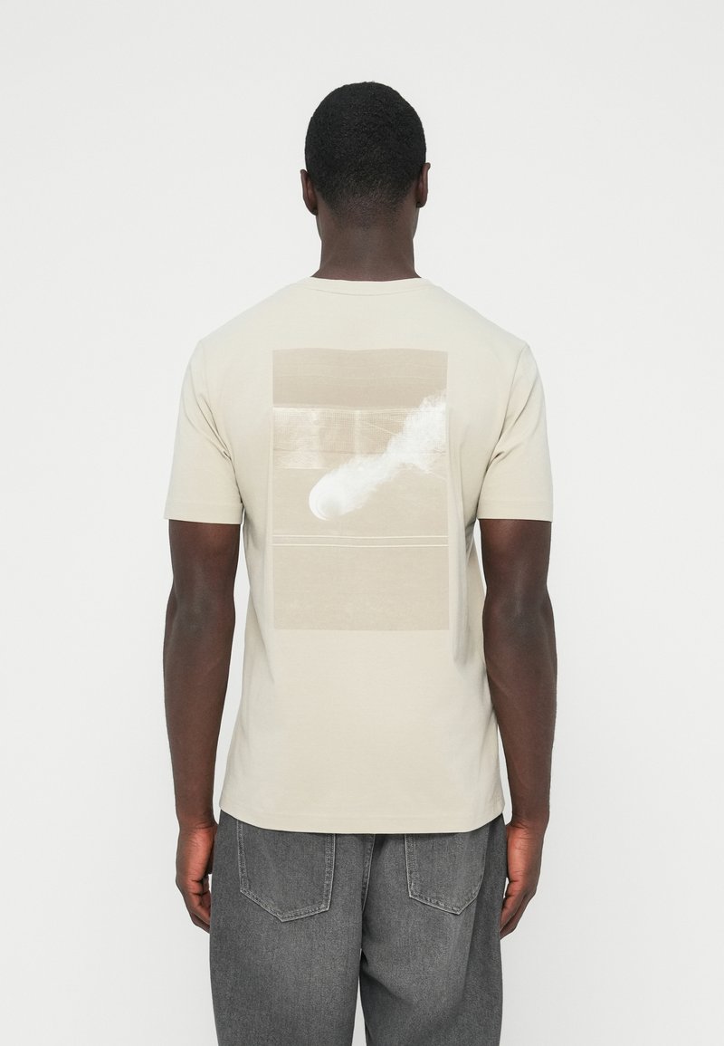 Béžna bombažna majica s kratkimi rokavi, na zadnji strani z otipljivim grafičnim potiskom v nevtralnih odtenkih. Gladek material, sproščen kroj.