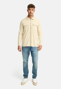 Ung mand står iført en beige zip-up jakke, lyseblå jeans og hvide sneakers på en helt hvid baggrund.