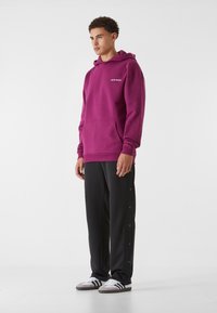 Felpa viola con cappuccio e tasca a marsupio, pantaloni sportivi neri con bottoni laterali, e sneaker bianche con strisce nere.