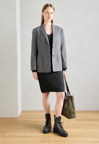Blazer de mezcla de lana en gris, vestido de punto acanalado negro, botas negras con cordones y herrajes dorados, bolso tote de ante verde, de pie sobre un suelo de madera.