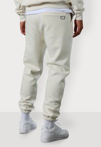 Hellbeige Jogginghosen mit figurbetontem Design, elastischen Bündchen und einem kleinen schwarzen Logo-Detail an der Gesäßtasche. Kombiniert mit weißen Sneakern.
