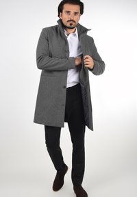 Abrigo de mezcla de lana gris con un cuello alto, cierre de botones y bolsillos laterales. Usado sobre una camisa blanca y combinado con pantalones oscuros y zapatos marrones.