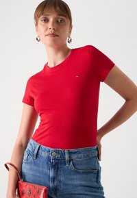 T-shirt rouge à manches courtes en coton avec un col rond et un petit logo sur la poitrine, assorti à un short en jean bleu clair et un sac à motif.