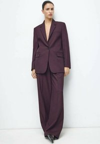 Bordowy, szyty na miarę garnitur z marynarką na klapach z nacięciem i szerokimi spodniami, oba wykonane z teksturowanej tkaniny z dwoma przednimi kieszeniami.