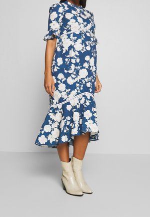 Zwangere persoon draagt een blauwe midi-jurk met bloemenprint en een gerimpelde zoom en crème enkellaarsjes, staand op een grijze vloer tegen een eenvoudige muur.
