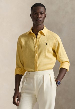 Żółta lniana koszula z kołnierzykiem zapinanym na guziki i małym haftowanym logo. Noszona z białymi spodniami z wysokim stanem i zegarkiem na nadgarstek.