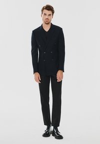 Granatowa marynarka dwurzędowa z wyłogami, połączona z czarną koszulą typu V-neck i dopasowanymi czarnymi spodniami, wykończona błyszczącymi czarnymi butami.