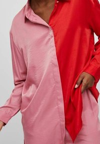 Camicia bicolore con lato sinistro di un rosa tenue e lato destro di un rosso vivace, realizzata in tessuto liscio con colletto a bottoni e maniche lunghe.