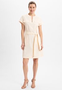 Frau steht lächelnd da, trägt ein hellbeiges, kurzärmeliges Kleid mit Taillengürtel und hautfarbene High-Heel-Sandalen vor weißem Hintergrund.