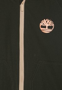 Giacca verde scuro con cerniera e finitura testurizzata, caratterizzata da una cerniera beige e un logo di un albero in rame sul lato sinistro. Sono visibili due tasche laterali.