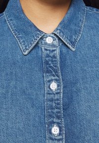 Gros plan d’une personne portant une chemise en jean bleue avec trois boutons blancs fermés à l’avant et un col pointu.