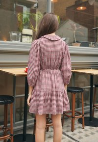 Red and white gingham dress with a collar, fitted waist, and tiered skirt. Set in a café with wooden furniture and indoor plants visible.