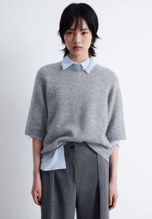 Grau entspannter Pullover aus weichem Material, über einem hellblauen Kragenshirt getragen. Das Outfit beinhaltet lockere graue Hosen mit Falten.