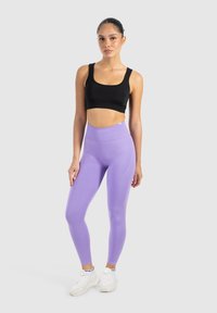 Czarny top crop i lawendowe legginsy o wysokim stanie z elastycznego materiału. Modelka stoi w białych sneakersach, na tle neutralnym.