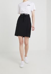 Jupe noire avec taille élastique et détail de cordon, assortie à un t-shirt blanc et des baskets blanches, se tenant sur un fond neutre.
