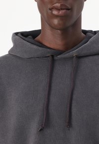 Felpa grigia con cappuccio realizzata in un tessuto testurizzato, dotata di un cordino con dettagli dorati. Vista ravvicinata che mette a fuoco il colletto e il cappuccio.