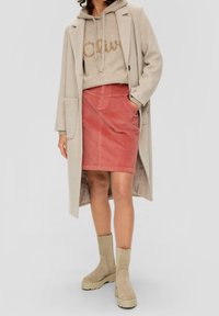 Femme portant un long manteau beige, un hoodie tan avec inscription, une jupe en velours côtelé rose, et des bottes plateformes beiges, se tenant avec une main dans la poche.