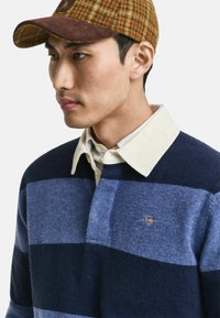 Blue and navy striped sweater with a white corduroy collar. Sports a small embroidered logo on the chest. Accompanied by a checkered cap.
