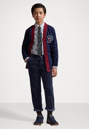 Cárdigan azul marino con ribete rojo, logo en forma de P, camisa blanca de botones con estampado, corbata a rayas verdes, pantalones de pana azul marino y zapatillas azules marinas con suelas en color marrón claro.