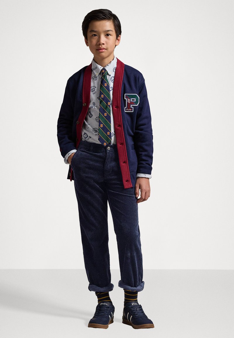 Marine cardigan met rode accentuering, P-logo, witte gebloemde button-up shirt, groen gestreepte das, marine corduroy pants en marine sneakers met beige zolen.