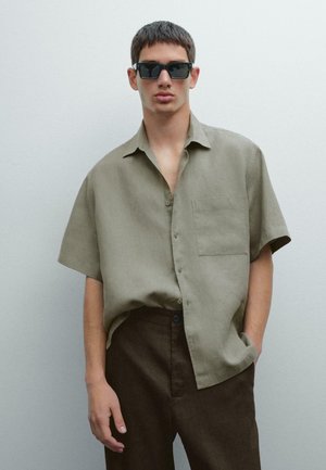 Short-sleeve olive-green linen shirt with a chest pocket and open collar, paired with brown trousers; model wears black sunglasses.