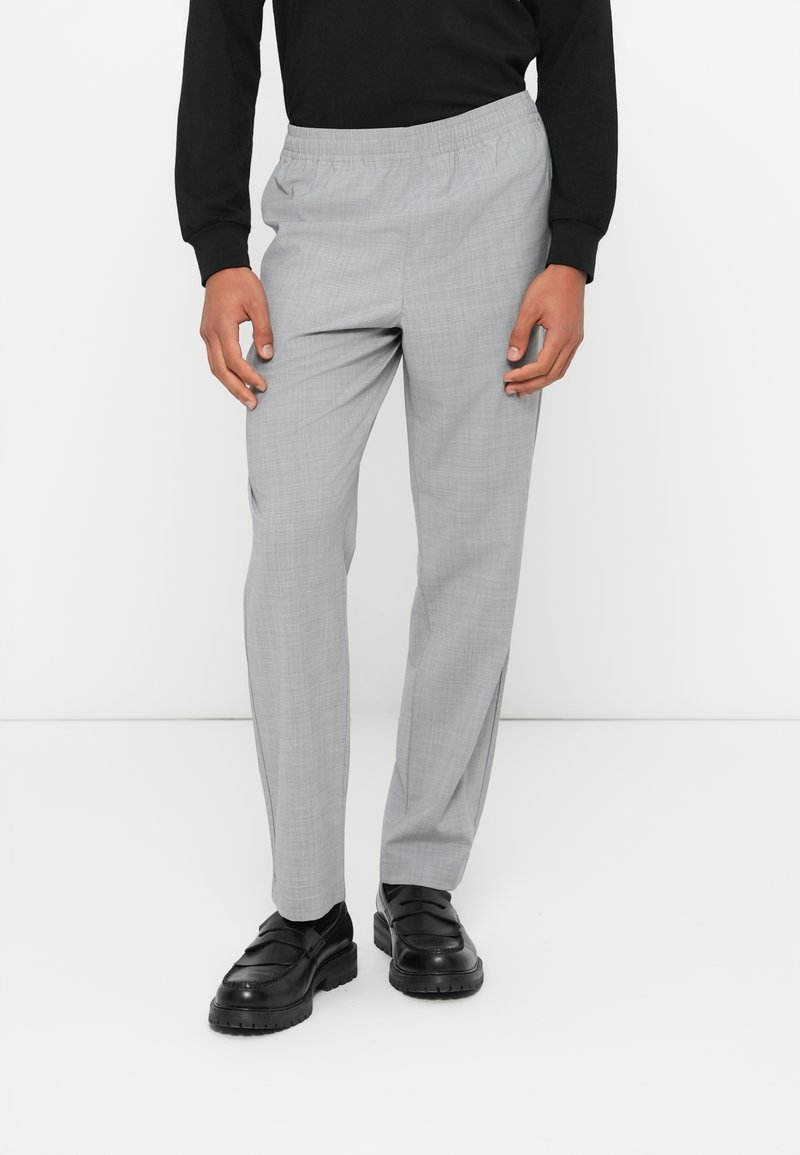 Pantalon gris clair avec un motif texturé, taille élastique et poches latérales, associé à des chaussures noires.