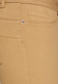 Los pantalones vaqueros de mezclilla color beige presentan una textura suave, costuras visibles y un pequeño bolsillo trasero con un acento de remache metálico.