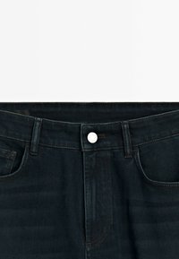 Jean en denim bleu foncé avec une coupe droite, dotée d'un bouton en métal blanc, de cinq poches et de surpiqûres contrastantes le long des coutures.