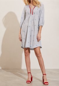 Jasnoniebieska bawełniana sukienka z haftowanymi kwiatowymi wzorami, czerwonymi akcentami wzdłuż dekoltu i rękawami 3/4 z bufkami. Nosi ją w połączeniu z czerwonymi sandałami na słupku.