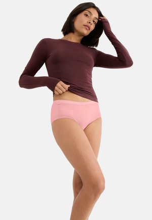 Frau hebt ein langärmliges bordeauxrotes Oberteil an, trägt hellrosa Hipster-Unterwäsche und steht vor einem schlichten hellen Hintergrund.