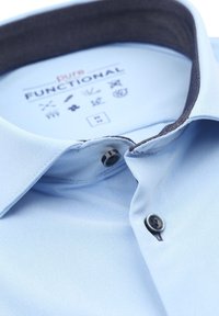 Lichtblauhemd met een donkere kraag, voorzien van een knoopsluiting. De stof heeft een gestructureerde weving, gelabeld "pure FUNCTIONAL" met verzorgingssymbolen.