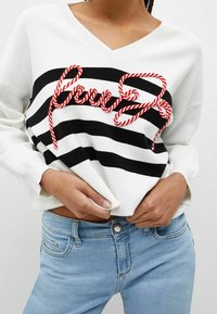 Pull en maille blanc avec des rayures horizontales noires et un texte brodé en rouge. Col en V, manches longues, assorti à un jean bleu clair.