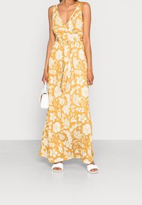 Robe maxi florale jaune avec un décolleté en V profond, design enroulé, taille cintrée et jupe fluide ; associée à des sandales blanches et un sac à main.