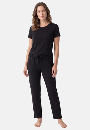Vrouw staat blootsvoets, draagt een zwarte top met korte mouwen en zwarte broek met koordjes tegen een egale lichte achtergrond.
