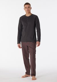 Junger Mann, der barfuß steht und ein schwarzes langärmliges Henley-Shirt sowie lockere gestreifte Schlafanzughosen trägt, vor einem weißen Hintergrund.