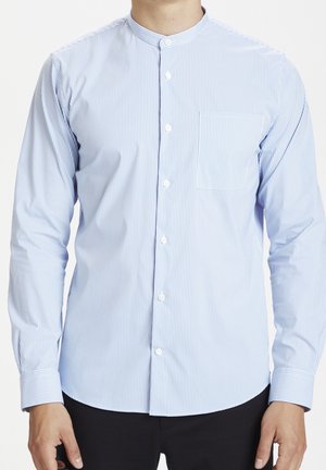 Mężczyzna ubrany w jasnoniebieską i białą cienko paskowaną koszulę z długim rękawem, z kołnierzem typu band collar i guzikami z przodu, w połączeniu z czarnymi spodniami.