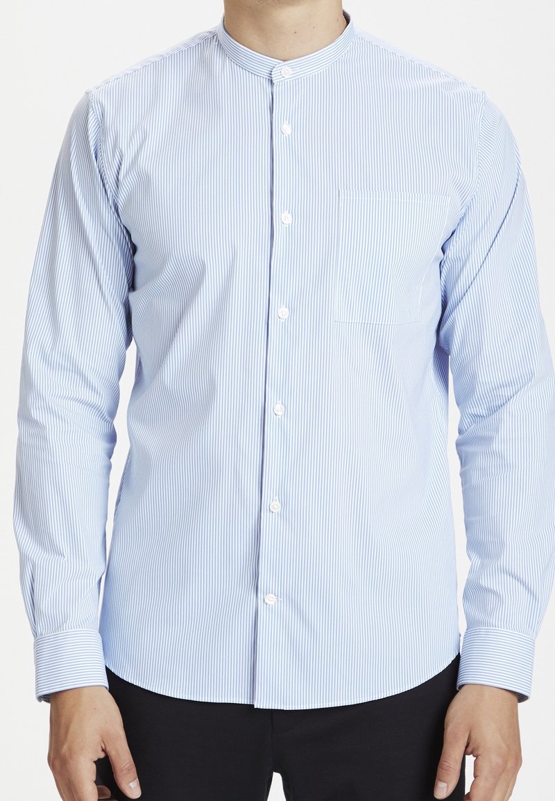 Homme portant une chemise à fines rayures bleu clair et blanc, à manches longues, avec un col mao et des boutons sur le devant, associée à un pantalon noir.