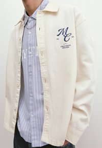 Light beige button-up shirt with a collar, featuring a navy print "MC" logo and text. Underneath, a light blue striped shirt is visible.