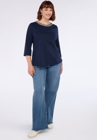 Frau steht da, trägt ein marineblaues Oberteil mit Kettendetail am Ausschnitt, weite blaue Jeans und weiße Sneaker vor einem schlichten weißen Hintergrund.