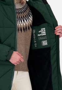 Grön quiltad jacka med svart fleecefoder, med en etikett som anger "PETA Godkänd Vegan" och tvättsymboler. Det inre mönstret framhäver hållbarhet.