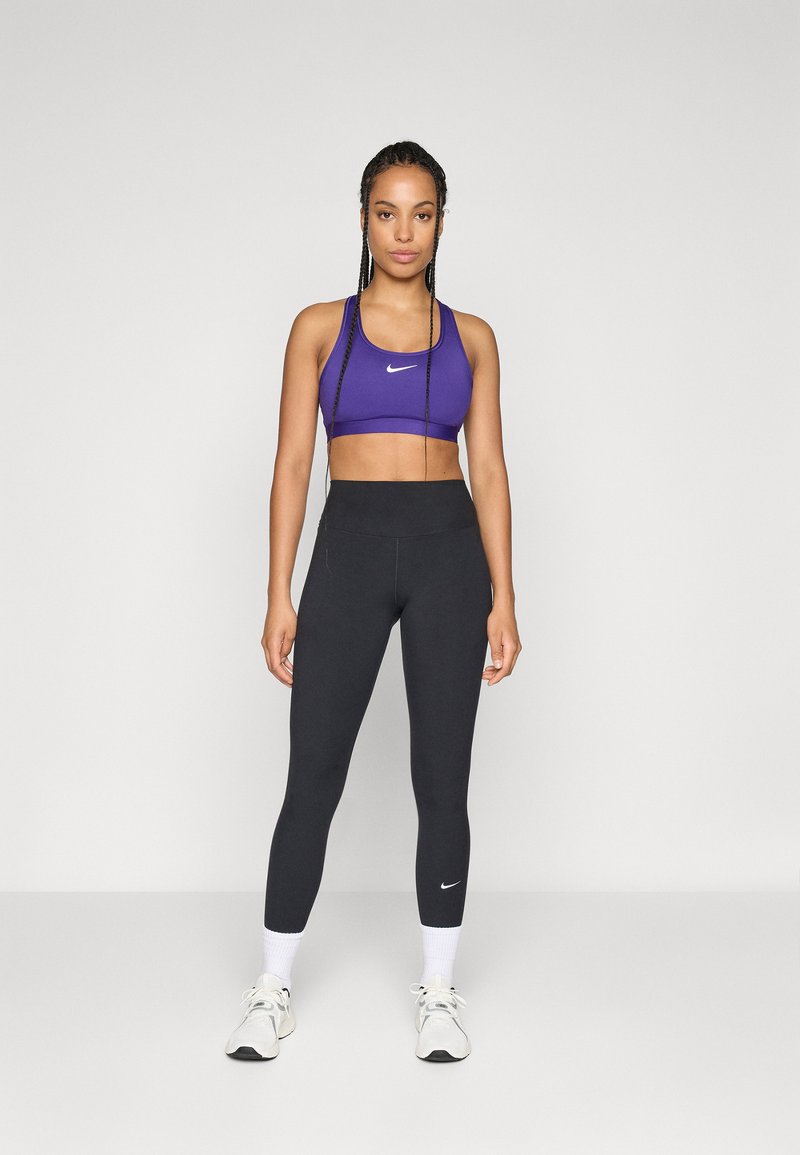 Femme debout portant un soutien-gorge de sport violet, un legging noir taille haute, des chaussettes blanches et des baskets blanches sur un fond uni.