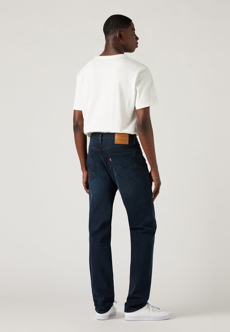 Jeans de mezclilla oscura con corte recto, parche de cuero en la cintura y camiseta blanca. Modelo mostrado de espaldas con zapatillas blancas.