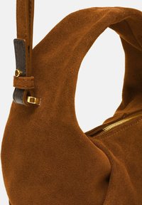 Bruine suede schoudertas met een zachte textuur, gebogen vorm, gouden rits en leren strapaccenten. Beschikt over een gestructureerde opening.