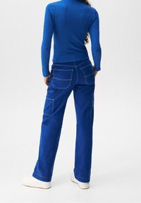 Haut à manches longues bleu texturé associé à un pantalon cargo ample bleu avec poches arrière et coutures contrastées, porté avec des baskets blanches.