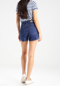 Pantalones cortos de mezclilla de cintura alta en azul oscuro con un dobladillo deshilachado, que cuentan con dos bolsillos traseros y un diseño ajustado. Combinados con una blusa a rayas y zapatos casuales.